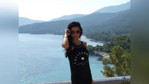 Young woman in a black California graphic tank top relaxing by a scenic turquoise ocean coastline.