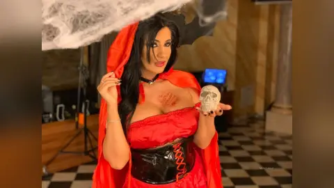 MissyJolie poses in a red hooded cape and black corset, holding a white skull near her chest.