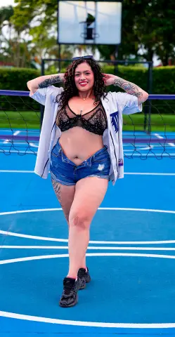 Smiling tattooed woman in a white jersey, black lace bra, and denim shorts posing on a blue outdoor basketball court.
