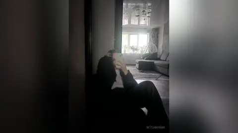 RittaMyers holds a phone to her face while sitting on a dark floor in a bright living room.
