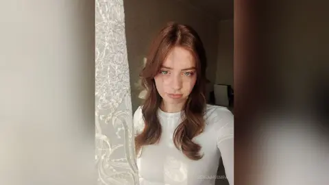 Young woman with wavy brown hair wearing a white long-sleeve shirt in a bright, minimalist indoor setting.