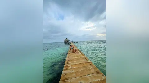 Woman in a bikini relaxing on a wooden pier over turquoise ocean water under a cloudy sky.
