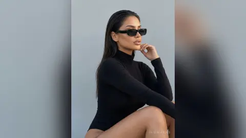 Stylish woman in a black turtleneck and sunglasses posing against a minimalist grey background.