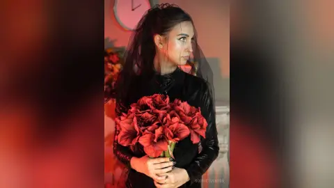 Elegant woman in a black gothic dress holding a large bouquet of red flowers in a moody, dark setting.