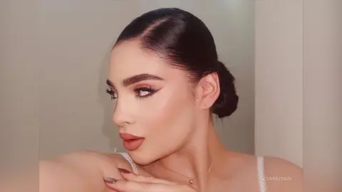 Woman with sleek dark hair and glamorous winged eyeliner wearing a white lace top in a soft, elegant setting.