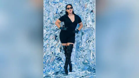 KimRoberth stands in a black bodysuit and black gloves against a shiny silver tinfoil background.