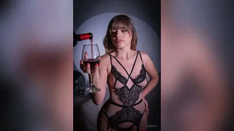 Young woman in black lace lingerie holding a glass of red wine in a moody, dimly lit setting.