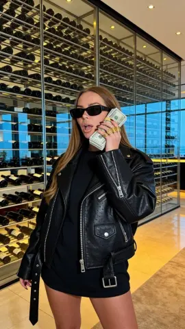 Stylish woman in a black leather jacket holding cash in a modern luxury wine cellar.