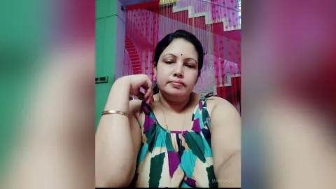 ManishaJi — ManishaJi rests her chin on her hand while wearing a colorful patterned tank top in front of red curtains.