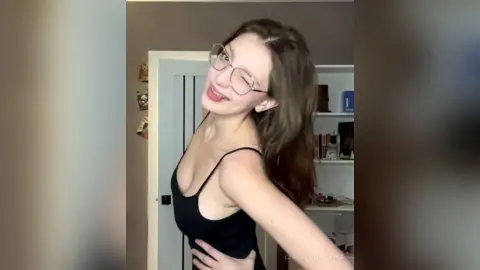 Smiling young woman with glasses in a black tank top posing playfully in a bright indoor setting.
