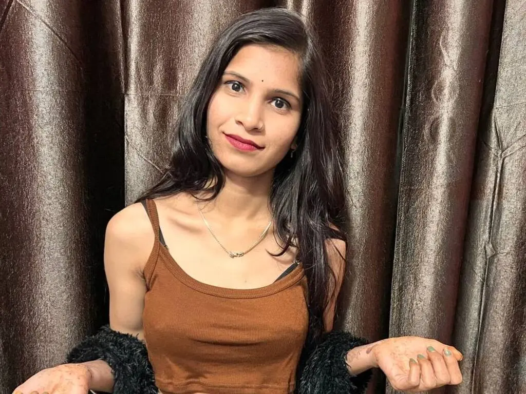 Young woman in a brown ribbed tank top posing against brown curtains in a cozy indoor setting.
