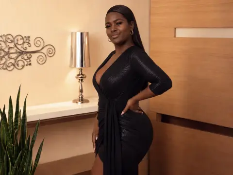 Smiling Black woman in a chic black evening dress posing in a warm, modern living room setting.