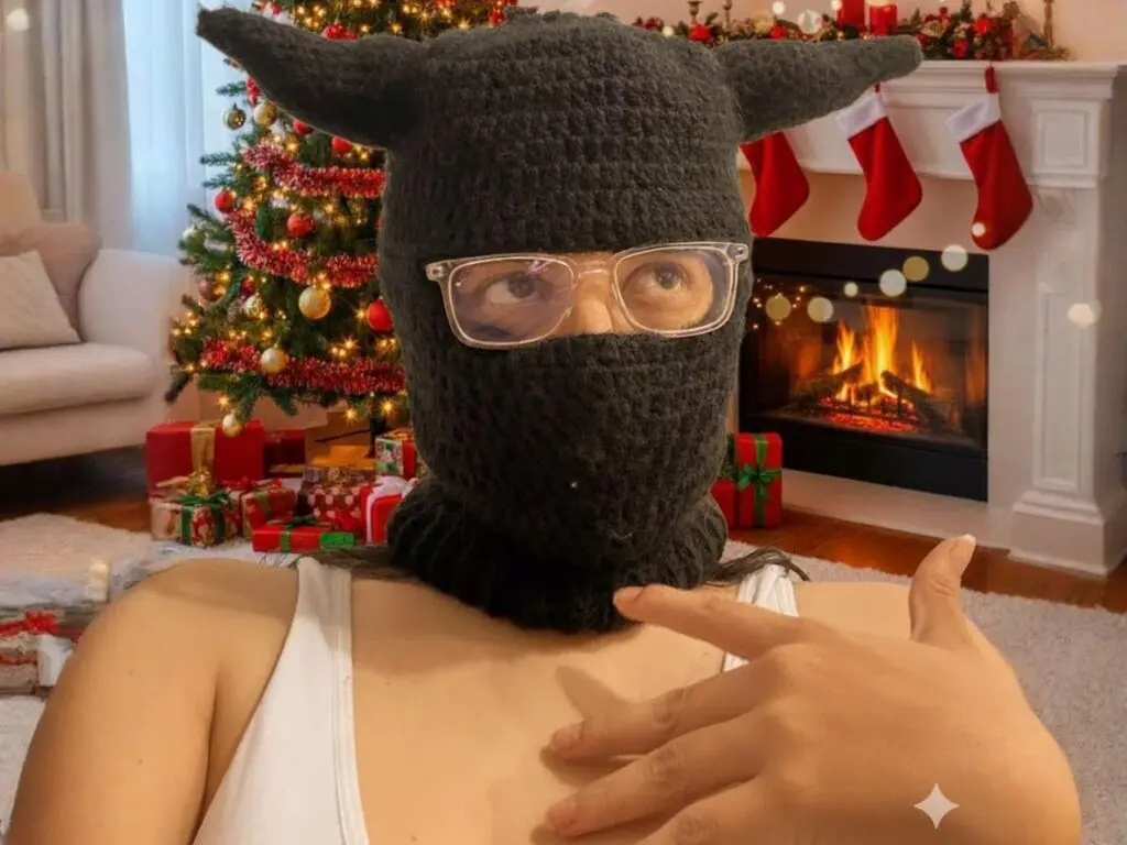 Woman in white tank top wearing a dark grey crochet devil horn hat sitting by a cozy lit Christmas tree fireplace.