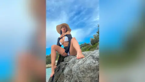NicoleBella sits on a large rock by the ocean wearing a white bikini top and a black mesh cover-up.