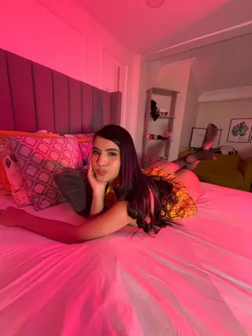 Young woman in a plaid outfit relaxing on a bed under warm pink lighting in a cozy bedroom.