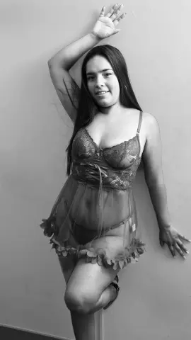 Smiling young woman in a lace lingerie bodysuit and sheer ruffled skirt posing against a plain wall.