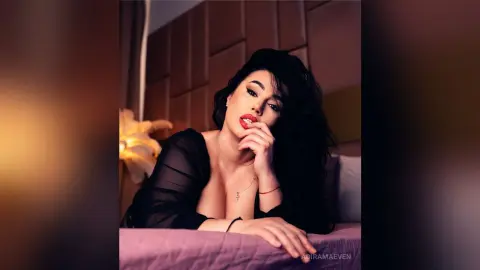 AdiraMaeven poses with long black hair and red lipstick, wearing a black sheer top while resting her chin on her hand.
