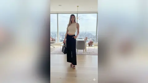 Stylish woman in a white tank top and black wide-leg trousers standing in a bright modern luxury apartment.