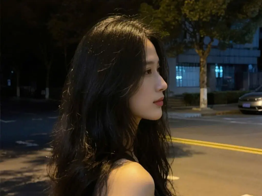 Young woman with long wavy black hair posing outdoors at night in a casual urban setting.
