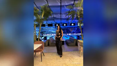 Stylish woman in a black bodysuit and trousers standing in a modern blue-lit lounge with palm trees.