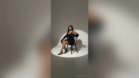 Elegant woman in a chic black cocktail dress and gloves sitting on a chair in a minimalist studio setting.