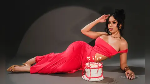 Elegant woman in a red evening gown reclining next to a birthday cake in a moody studio setting.