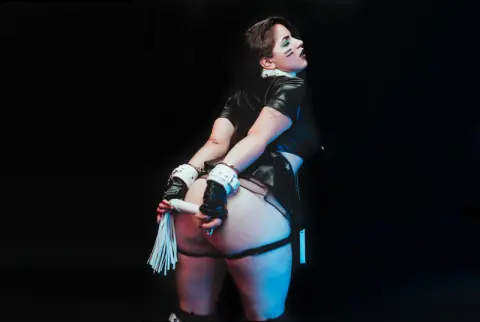 AbbyShinee poses facing away from the camera, showing her bare buttocks and wearing a black outfit with white wrist cuffs against a black background.
