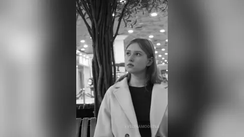 Melancholic young woman in a white coat standing by a tree in a dimly lit outdoor cafe setting.