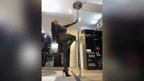 NicoleBella poses naked against a silver dance pole, wearing sheer black stockings and high heels.
