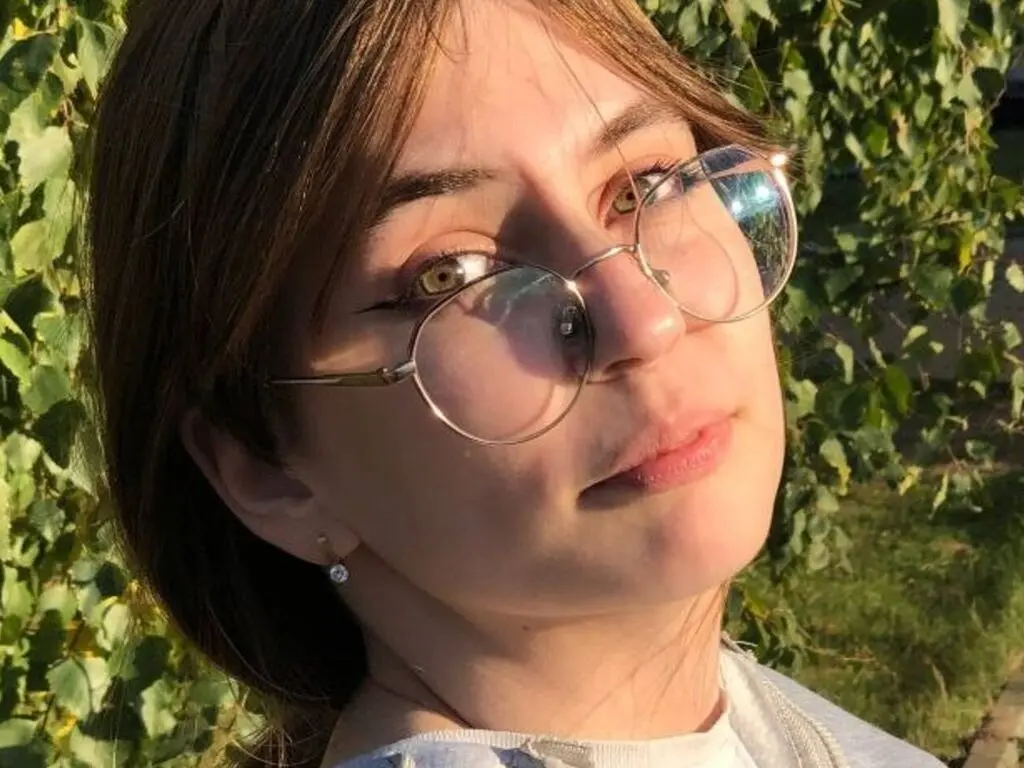 Smiling young woman with glasses in warm sunlight against a lush green garden background.