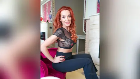 Smiling redhead woman in a black lace crop top and trousers sitting in a bright modern living room.