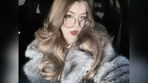Young woman with long wavy hair wearing round glasses and a grey faux fur coat sitting in a car.