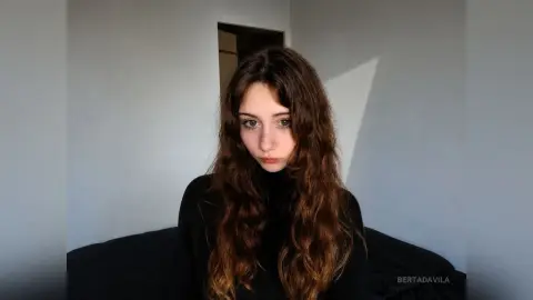 Young woman with long wavy brown hair wearing a black turtleneck in a minimalist room with soft lighting.