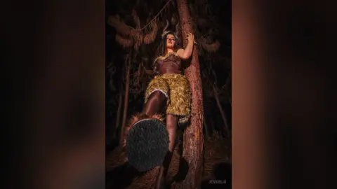 JessieLiu poses in a brown furry costume with pointed ears, leaning against a tree trunk in a dark forest setting.