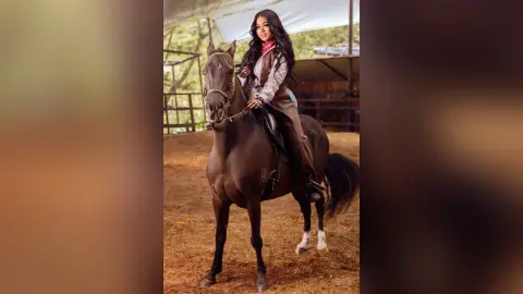 Elegant woman in equestrian attire riding a brown horse in a rustic stable setting.