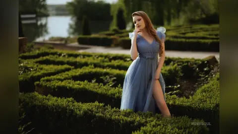 RubySoul poses in a long, light blue dress with a high slit, standing in a lush green hedge maze near a lake.
