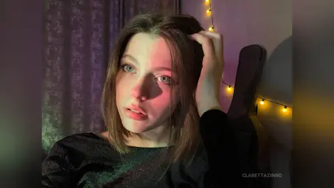Young woman in a black top sitting in a dimly lit room with warm fairy lights and a moody atmosphere.