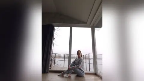 Young woman in a grey sweater and jeans sitting by a large window overlooking a serene lake view.