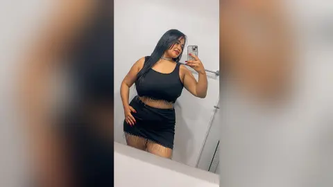 NiaCorelli poses in a black tank top and matching skirt with silver fringe detail, holding her phone for a mirror selfie.