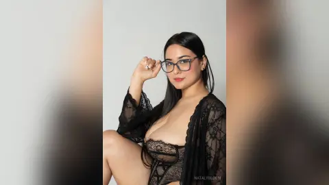Beautiful woman in black lace lingerie and glasses posing confidently against a plain white background.