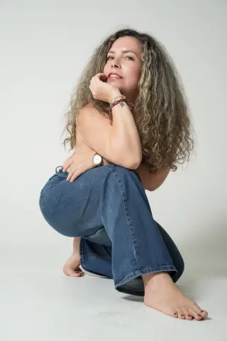 Smiling woman with long curly hair wearing blue jeans posing in a bright studio setting.