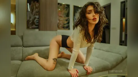Young woman in white long-sleeve crop top and black briefs posing on a grey sofa in a modern living room.