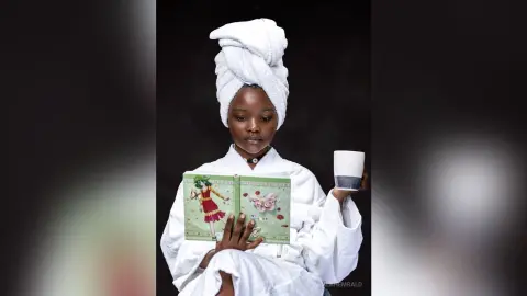 Relaxed Black woman in a white bathrobe and towel turban reading a book and holding coffee in a cozy mood.