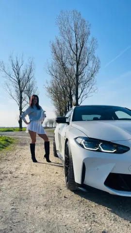 Stylish woman in a white skirt and black boots standing by a white luxury car on a sunny dirt path.