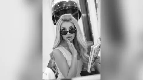 Stylish blonde woman in sunglasses holding a magazine in a moody black and white portrait.