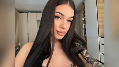 AriaVex poses with long black hair and heavy eye makeup, wearing a dark patterned top in a room with light grey cabinets.