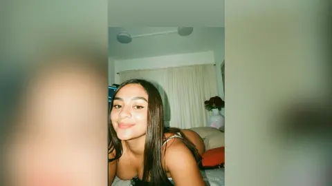 Young woman with long dark hair smiling in a cozy bedroom setting.