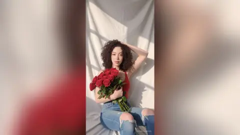 Young woman in a red tank top and blue jeans holding a bouquet of red roses in bright sunlight.