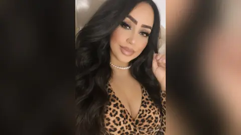 Beautiful woman with long wavy hair wearing a leopard print top and pearl necklace in a soft indoor setting.