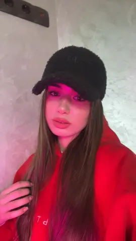 Young woman in a red hoodie and black fuzzy cap posing with a moody pink lighting aesthetic.